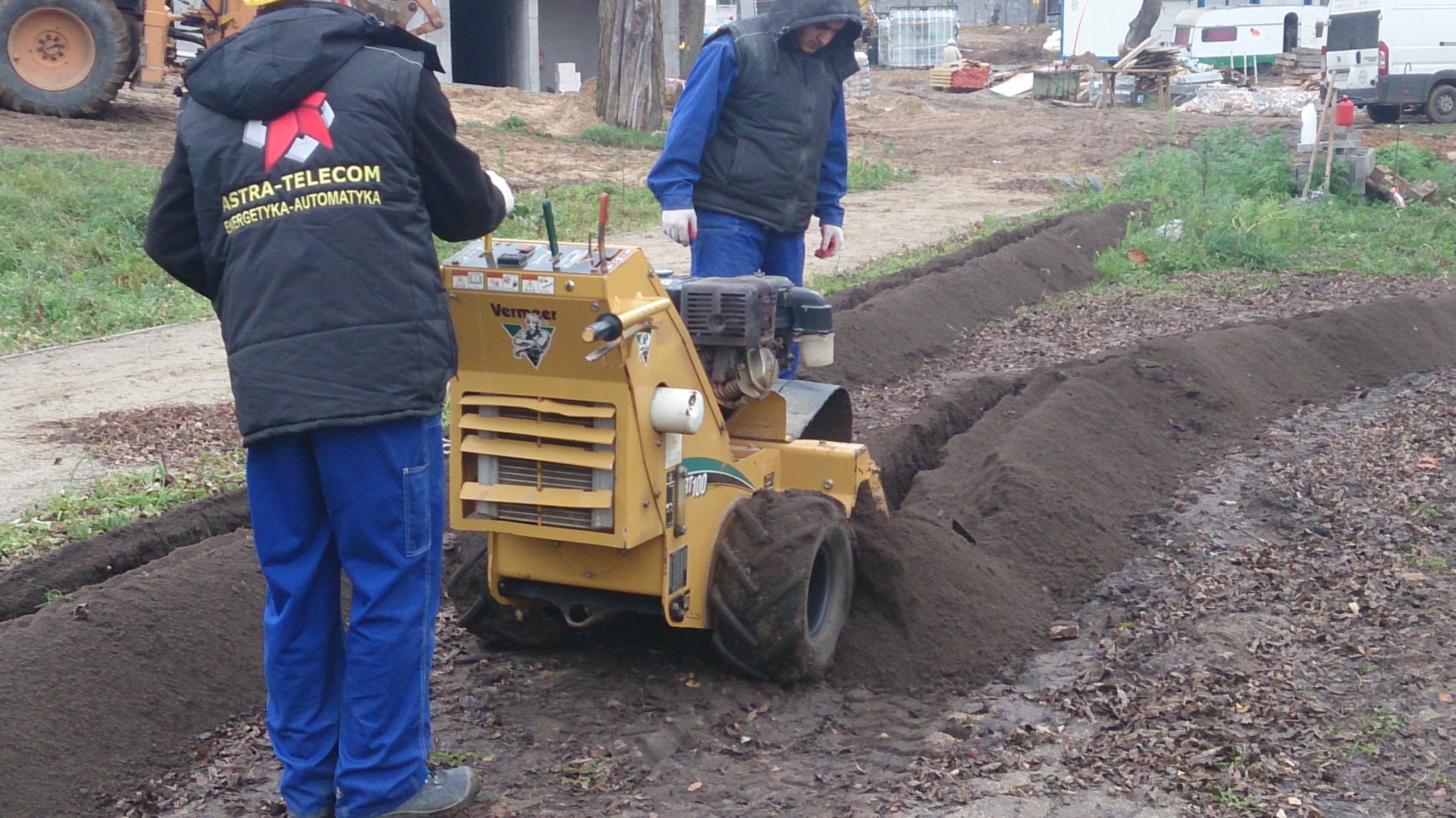 Żółta glebogryzarka Vermeer RT100 w trakcie wykonywania wykopu pod kable elektryczne, dwóch pracowników w odzieży roboczej nadzoruje prace, w tle teren budowy.