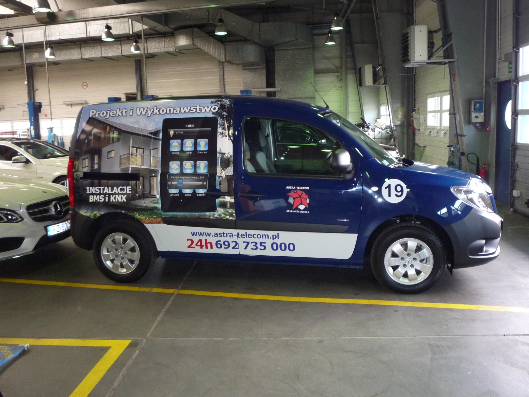 Niebieski samochód dostawczy z grafiką przedstawiającą dom z systemem inteligentnego sterowania oświetleniem, logo firmy Astra-Telecom i numerem telefonu, zaparkowany w warsztacie.