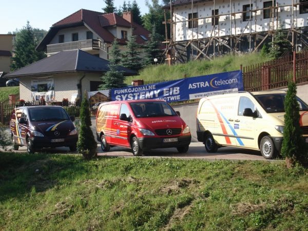 Trzy samochody dostawcze firmy Astra Telecom zaparkowane przed budynkiem w trakcie ocieplania, widoczny baner reklamowy firmy.
