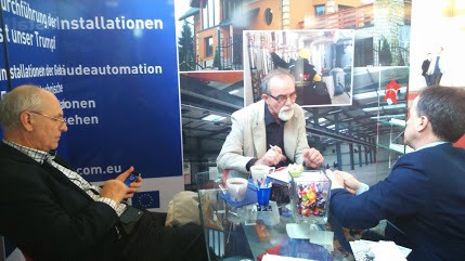 Trzech mężczyzn siedzi przy szklanym stole podczas rozmowy na tle plakatu prezentującego instalacje i budynek w trakcie prac. Jeden z mężczyzn trzyma telefon, drugi pisze, a trzeci słucha.