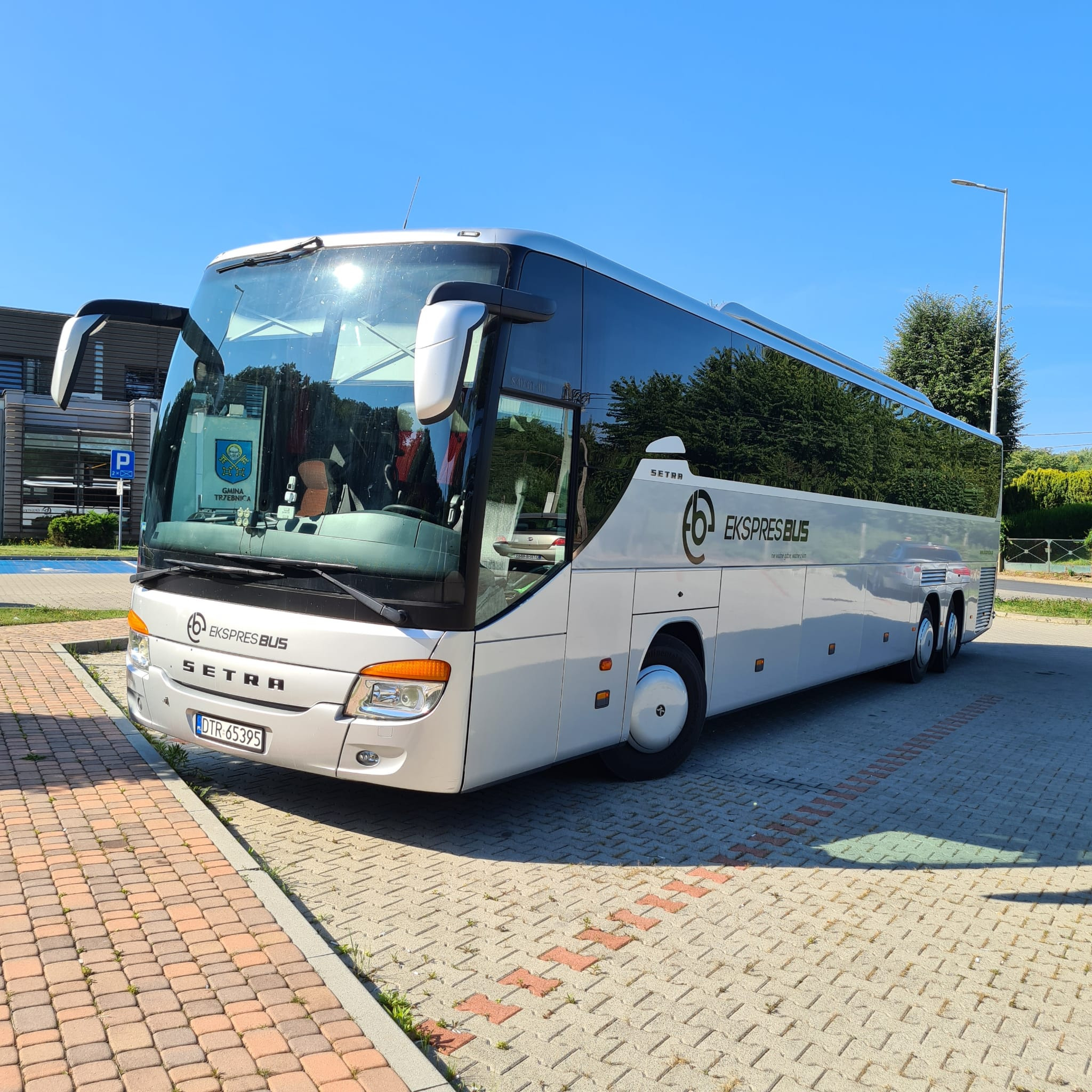 Biały autokar SETRA 417 'Ekspres Bus' zaparkowany na kostce brukowej, widoczny herb Gminy Trzebnica na przedniej szybie.