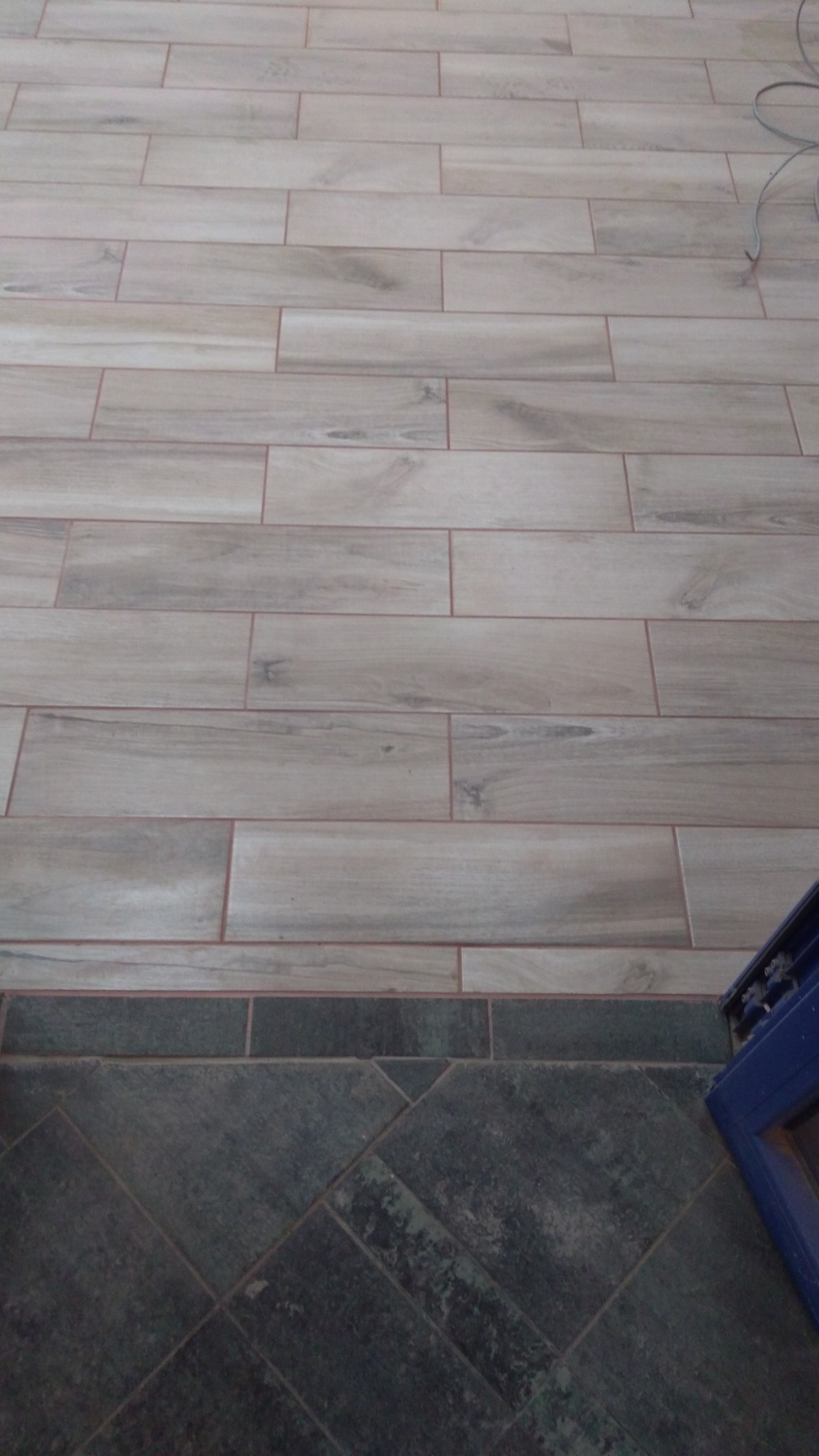 Ułożona podłoga z płytek imitujących drewno, przechodząca w ciemnozieloną, ułożoną w romby, z fragmentem niebieskiej ościeżnicy w prawym dolnym rogu.