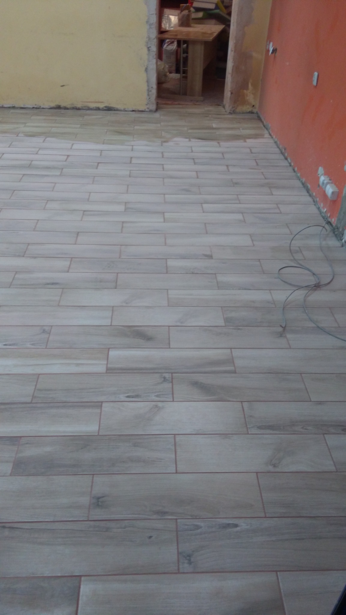 Ułożona podłoga z płytek imitujących drewno w odcieniach szarości, widoczne niedokończone ściany w kolorze żółtym i pomarańczowym, wystające kable elektryczne przy ścianie.