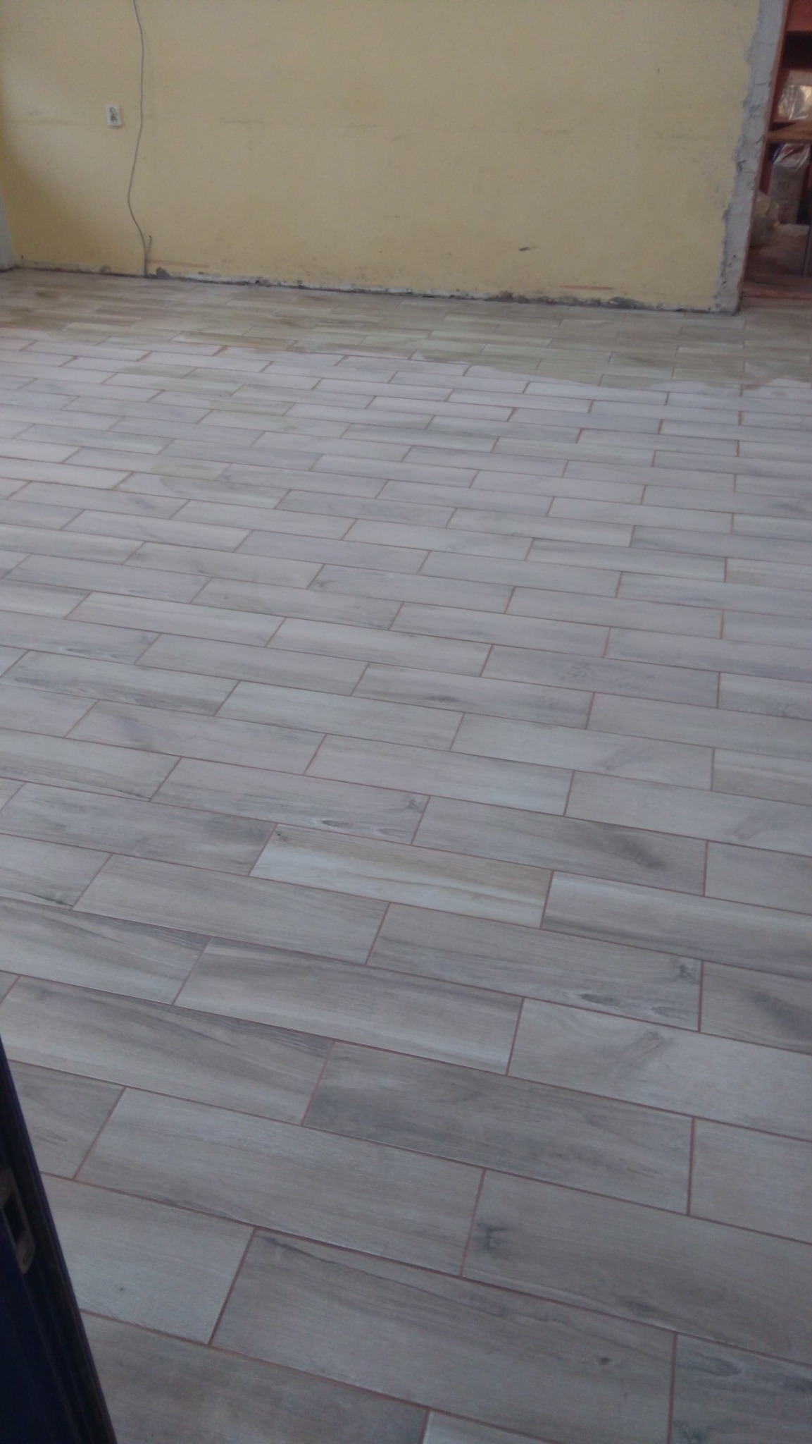 Ułożone płytki podłogowe imitujące drewno w pomieszczeniu w trakcie remontu, widoczne niedociągnięcia przy ścianie i ślady zaprawy.