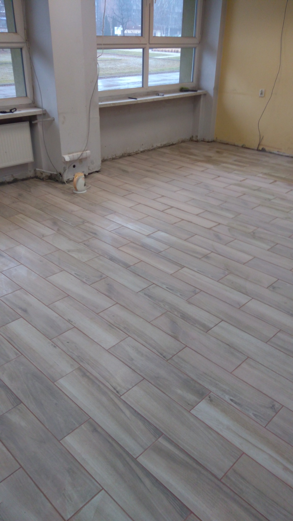 Ułożona podłoga z płytek imitujących drewno w jasnych odcieniach szarości, widoczne okna i ściany w stanie surowym, grzejnik i mały grzejnik elektryczny.