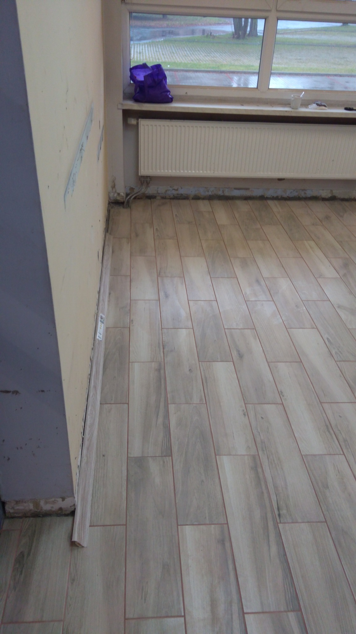 Ułożone płytki podłogowe imitujące drewno, widoczne niedokończone listwy przypodłogowe i ślady po pracach na ścianie, torba leży na parapecie okna.