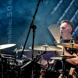 Perkusista w trakcie występu na scenie, uderzający w talerze z rozmytym ruchem pałeczek, widoczny napis 'mobile: 504' w tle.