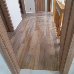 Położona pod kątem brązowa podłoga imitująca drewno, widoczne drzwi z drewnianą okleiną i fragment schodów z drewnianą balustradą.