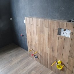CeraGress - Montaż stelaża podtynkowego WC na ścianie wyłożonej drewnopodobnymi, pionowymi płytkami; widoczne wystające rury instalacyjne z zaślepkami oraz ciemnoszare płytki na ścianie w tle.
