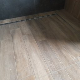 CeraGress - Ułożona podłoga z płytek imitujących drewno w kabinie prysznicowej z odpływem liniowym w Opolu. Ciemna ściana kabiny prysznicowej.