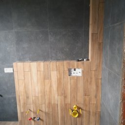 CeraGress - Montaż drewnopodobnych płytek na ścianie i podłodze w łazience, widoczne przygotowane instalacje wodne i elektryczne, szare płytki na ścianach w tle.