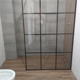 CeraGress - Kabina prysznicowa typu walk-in z czarnymi profilami w łazience z szarymi płytkami i drewnopodobną podłogą, widoczny odpływ liniowy i częściowo biała miska WC.