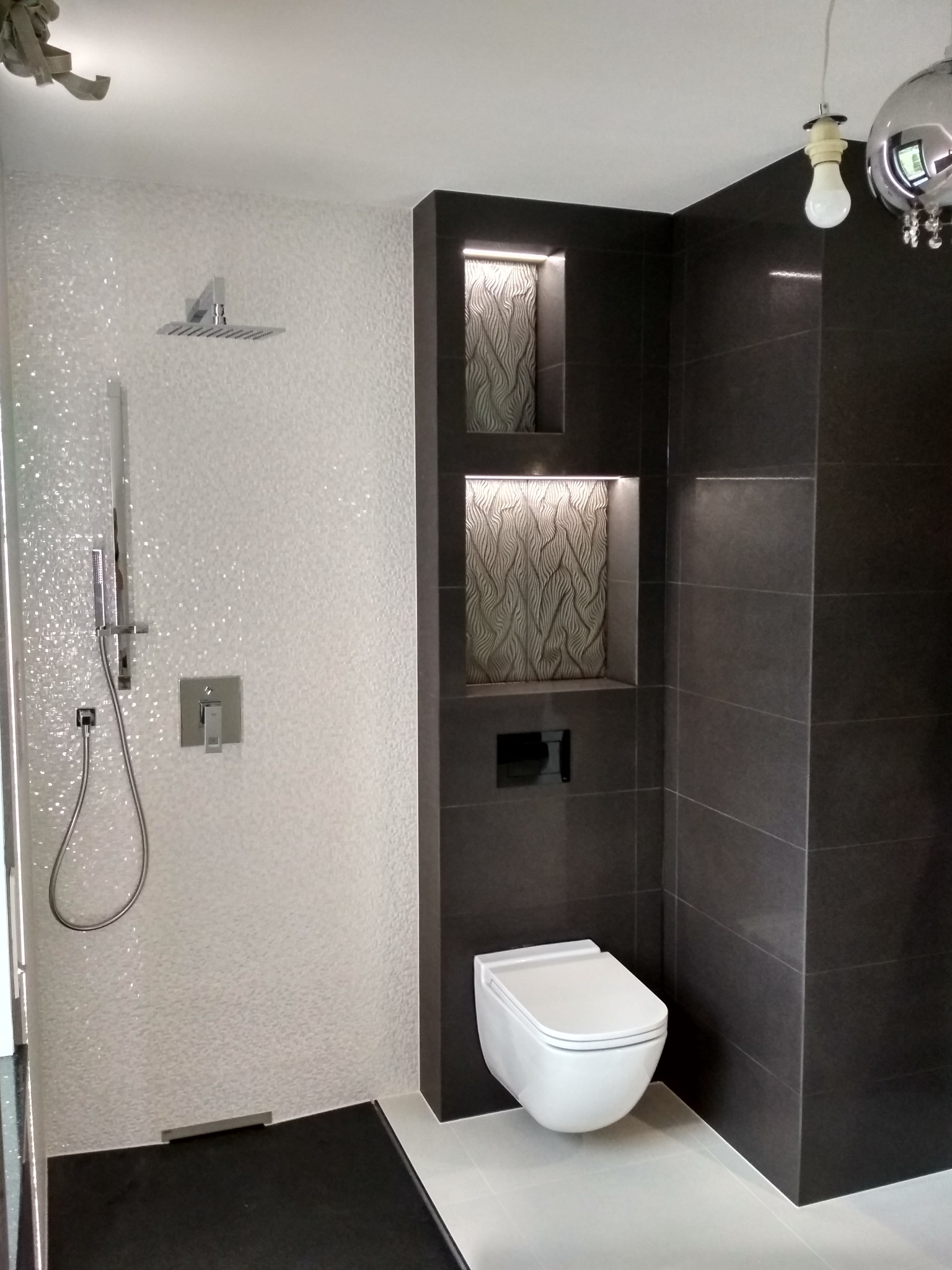 Nowoczesna łazienka z białymi, błyszczącymi płytkami pod prysznicem, ciemnoszarymi płytkami w strefie toalety, podświetlanymi wnękami z dekoracyjną teksturą i wiszącą miską WC.