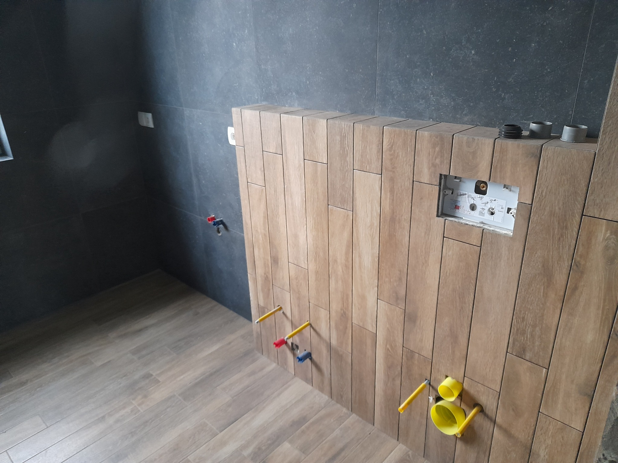 Montaż stelaża podtynkowego WC na ścianie wyłożonej drewnopodobnymi, pionowymi płytkami; widoczne wystające rury instalacyjne z zaślepkami oraz ciemnoszare płytki na ścianie w tle.