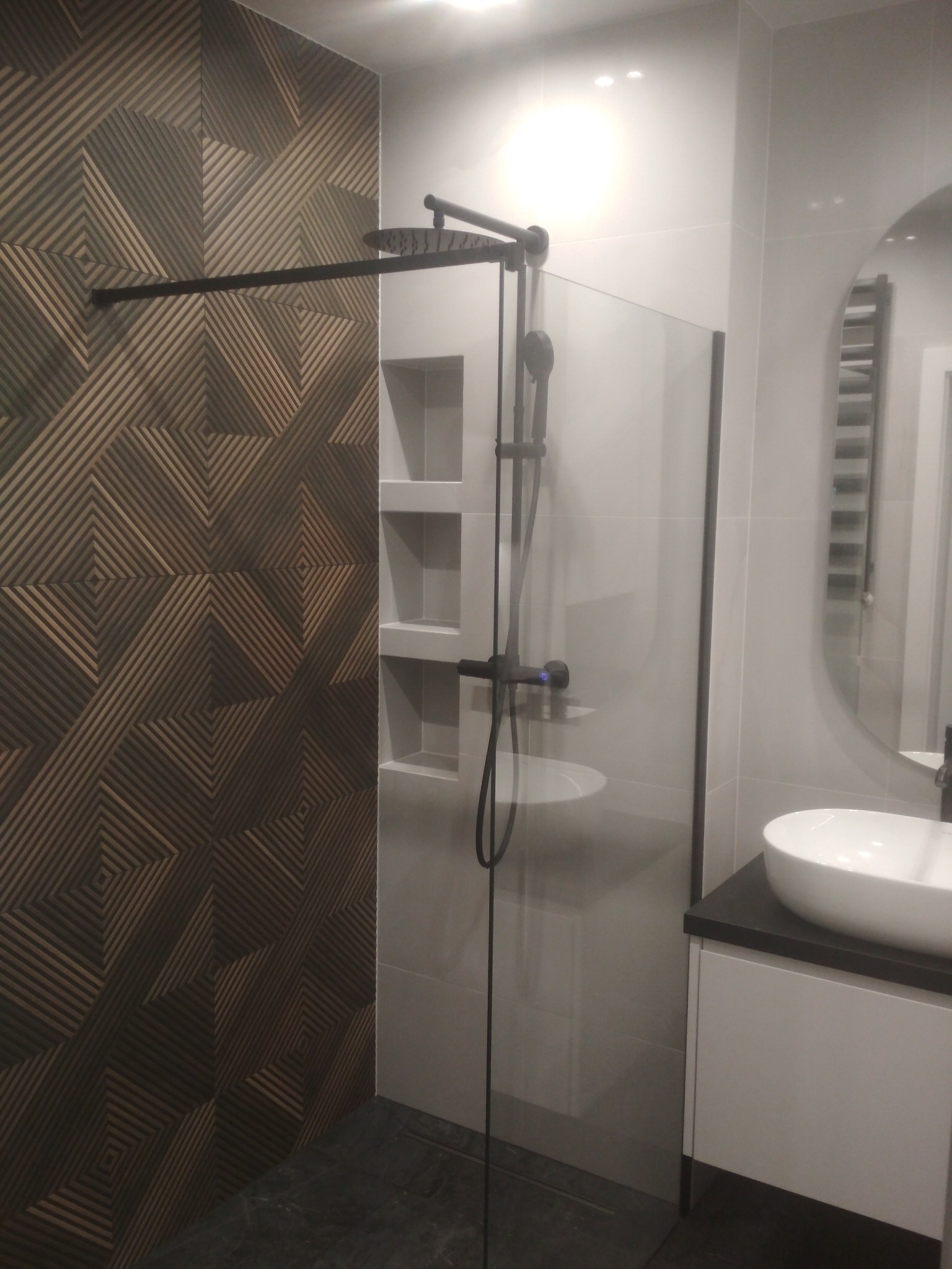 Nowoczesna łazienka z kabiną prysznicową typu walk-in, dekoracyjnymi płytkami na ścianie, wnękami półkowymi i owalną umywalką nablatową.