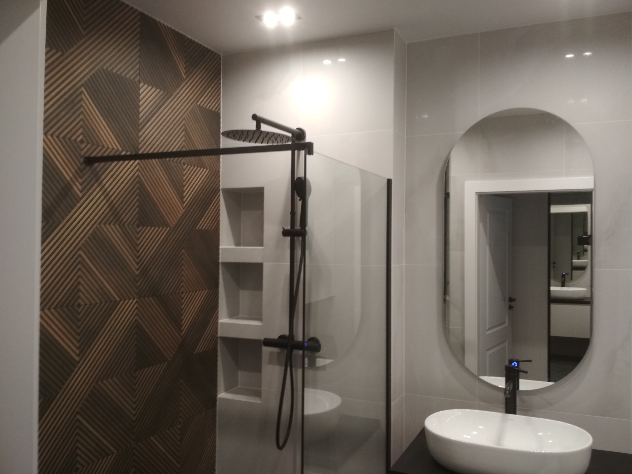 Nowoczesna łazienka z deszczownicą w czarnym kolorze, umywalką nablatową i ścianą z dekoracyjnymi panelami w geometryczny wzór w odcieniach brązu.
