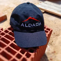 Granatowa czapka z daszkiem z wyhaftowanym logo firmy budowlanej ALDACH, leżąca na pustaku ceramicznym w plenerze.