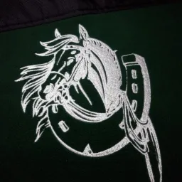 Biały haft przedstawiający głowę konia z elementami uprzęży na ciemnozielonym materiale, widoczny fragment czarnego materiału w górnej części