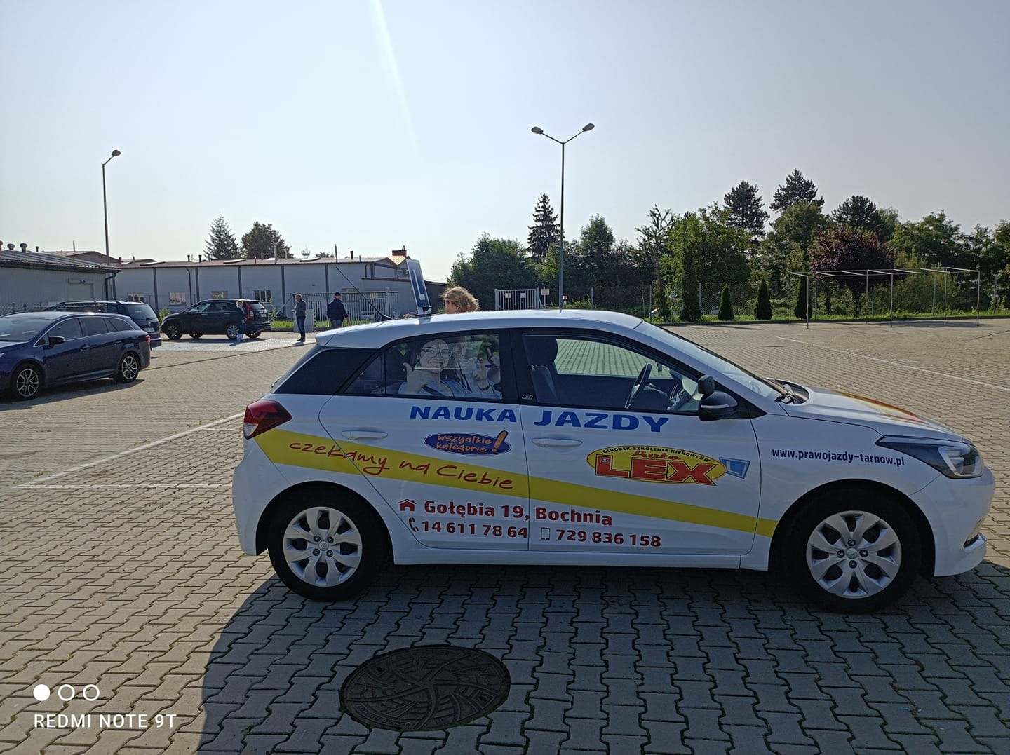 Biały samochód szkoły jazdy z napisem 'NAUKA JAZDY' i logo LEX na placu manewrowym, widoczne osoby w tle.