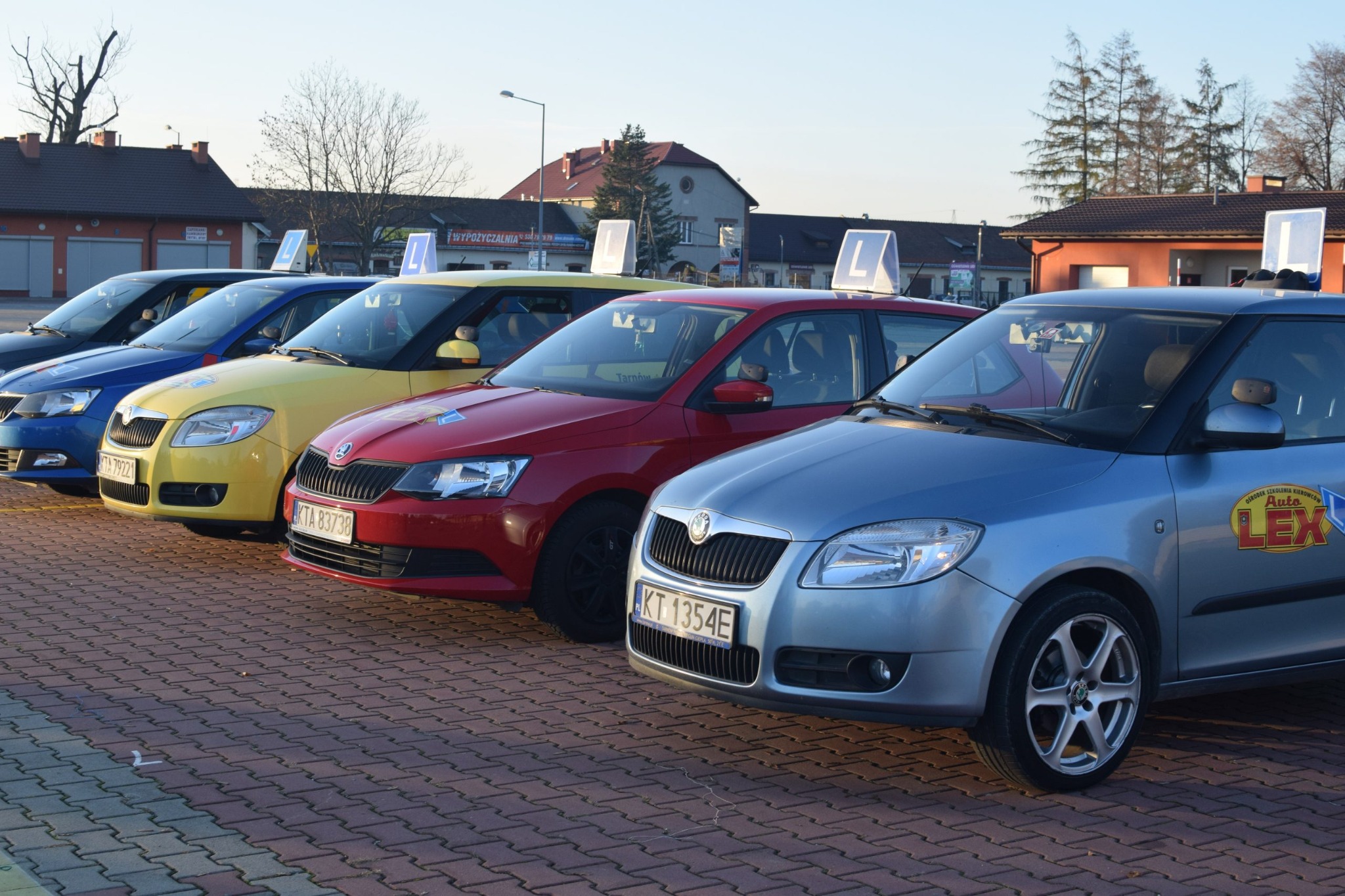 Flota samochodów Skoda Fabia w różnych kolorach (niebieski, żółty, czerwony, srebrny) z oznaczeniami 'L' na dachu, zaparkowana na placu manewrowym szkoły jazdy Auto Lex w Tarnowie.