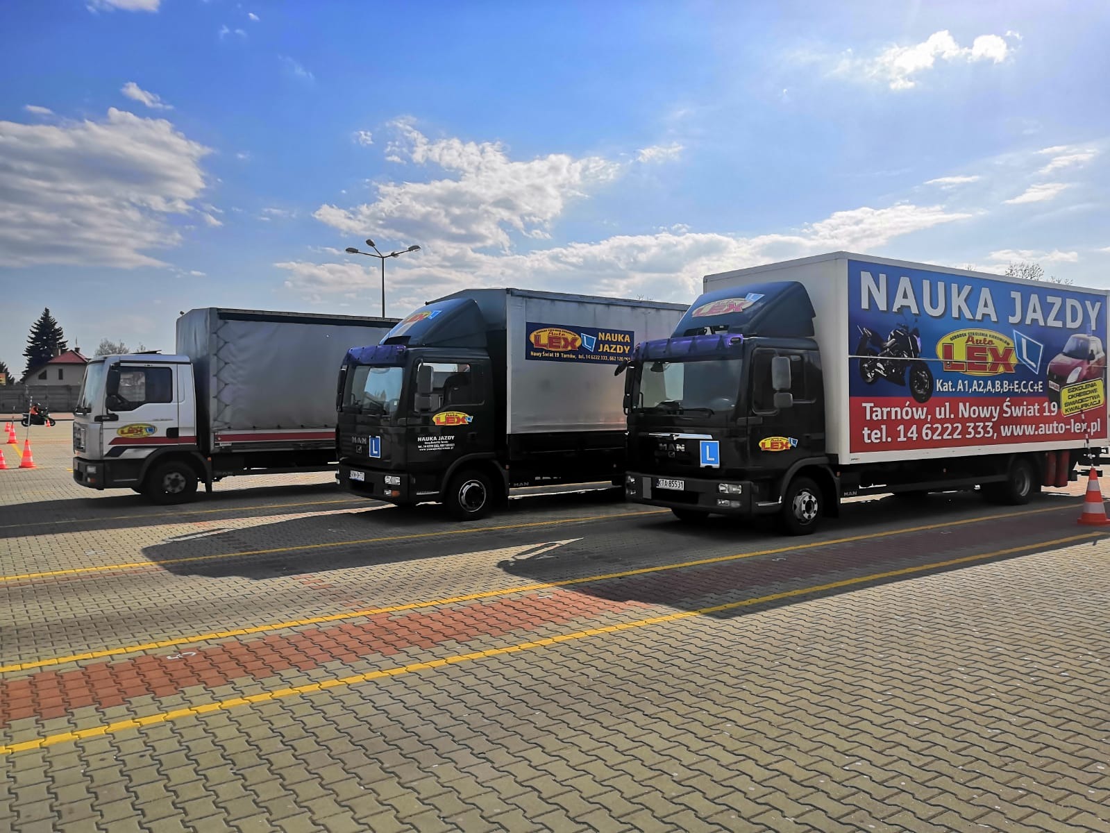 Trzy ciężarówki z oznaczeniami nauki jazdy zaparkowane na placu manewrowym z widocznymi pachołkami i oznakowaniem poziomym. Na burtach pojazdów reklama z informacjami kontaktowymi i kategoriami...