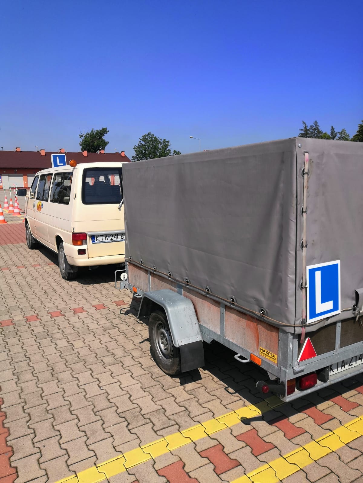 Beżowy bus z literą 'L' na dachu i przyczepa z szarą plandeką, również oznaczona literą 'L', stoją na placu manewrowym szkoły jazdy z kostki brukowej.