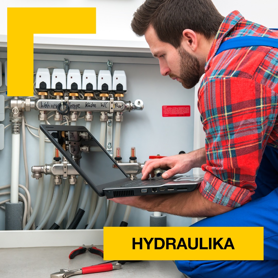 Hydraulik w czerwonej koszuli flanelowej, z brodą, konfiguruje system rozdzielaczy ogrzewania podłogowego za pomocą laptopa. Na podłodze leżą narzędzia hydrauliczne.
