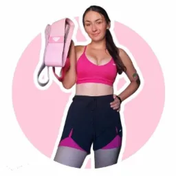 Uśmiechnięta kobieta w sportowym stroju, różowy top i czarne szorty z różowymi wstawkami, trzyma w dłoni różową taśmę do ćwiczeń na tle różowego okręgu.