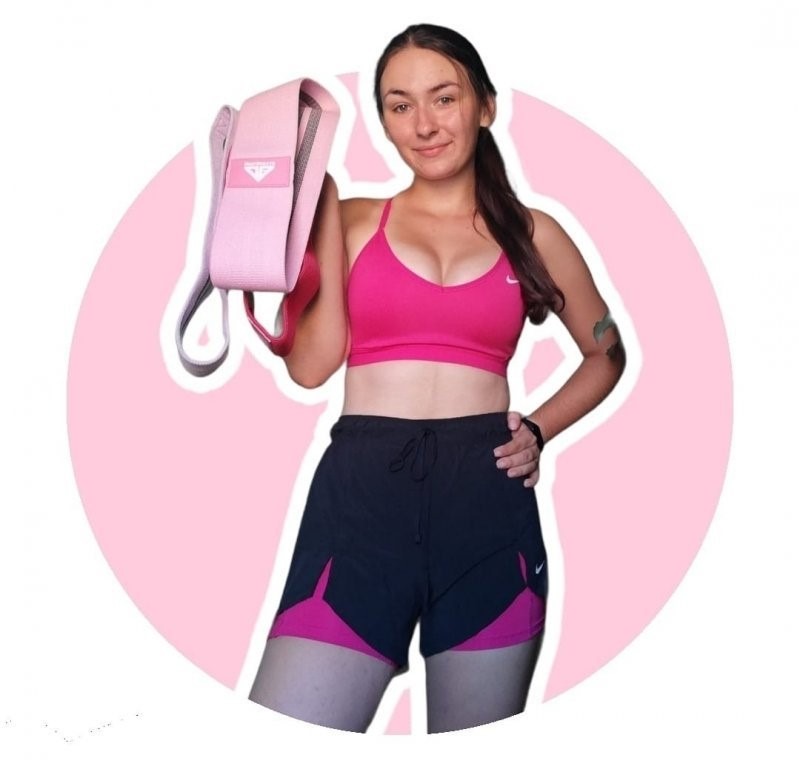 Uśmiechnięta kobieta w sportowym stroju, różowy top i czarne szorty z różowymi wstawkami, trzyma w dłoni różową taśmę do ćwiczeń na tle różowego okręgu.