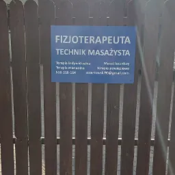 Tabliczka informacyjna 'Fizjoterapeuta Technik Masażysta' umieszczona na drewnianym płocie w Piastowie, z podanym numerem telefonu i adresem e-mail.