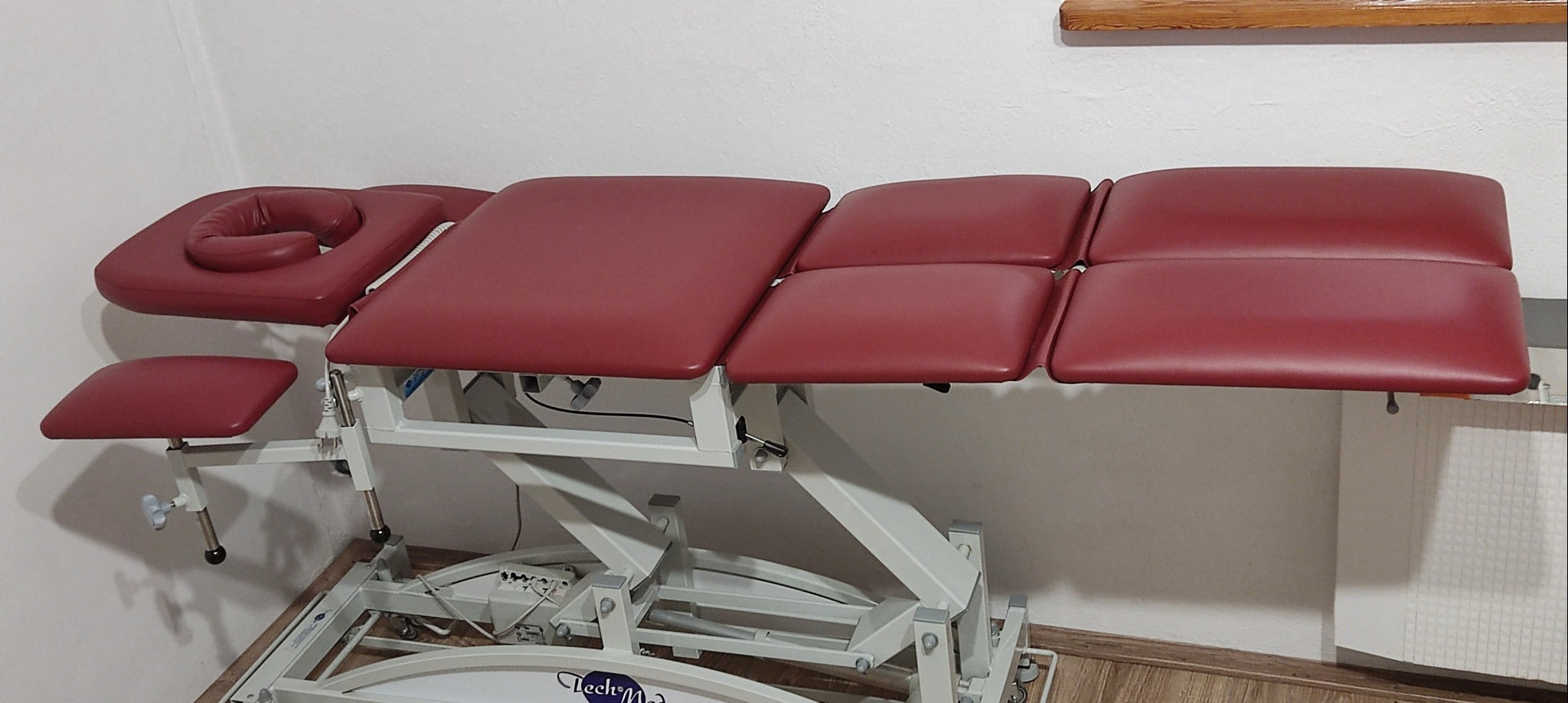 Rozłożony, regulowany stół do terapii manualnej z bordową tapicerką i białym stelażem, widoczny z boku, z zagłówkiem i dodatkowym podnóżkiem.