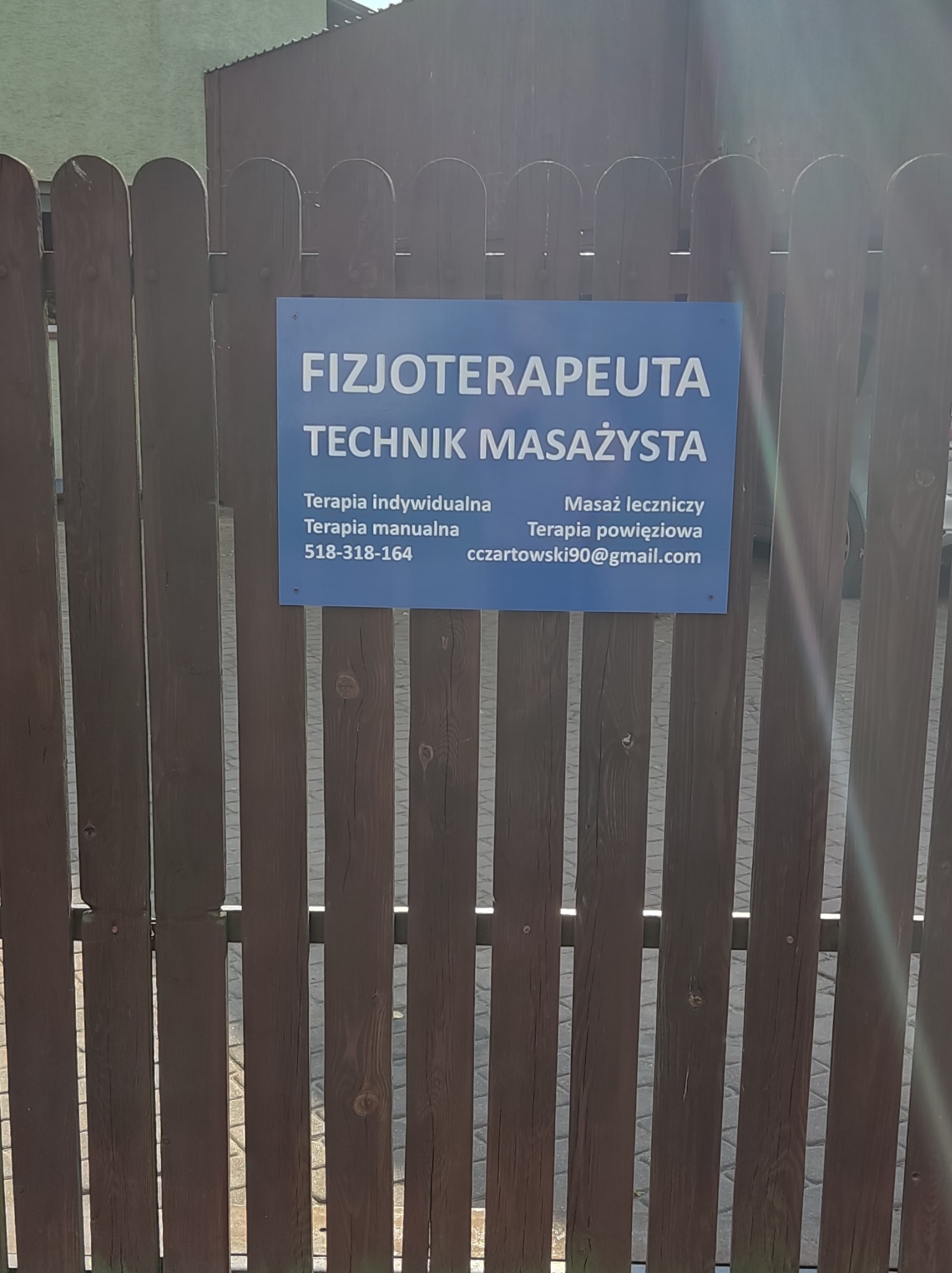 Tabliczka informacyjna 'Fizjoterapeuta Technik Masażysta' umieszczona na drewnianym płocie w Piastowie, z podanym numerem telefonu i adresem e-mail.