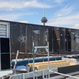 Centrala wentylacyjna z odzyskiem ciepła na dachu budynku, częściowo zabezpieczona folią, widok na wieżę telewizyjną i kościół w tle.