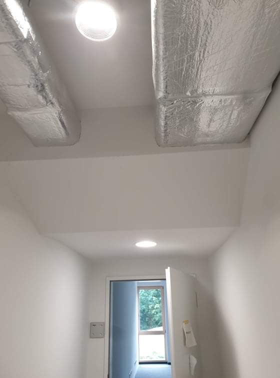 Widok z dołu na kanały wentylacyjne w izolacji aluminiowej, zamontowane pod sufitem w jasnym pomieszczeniu z widocznym oświetleniem punktowym i otwartymi drzwiami na końcu korytarza.