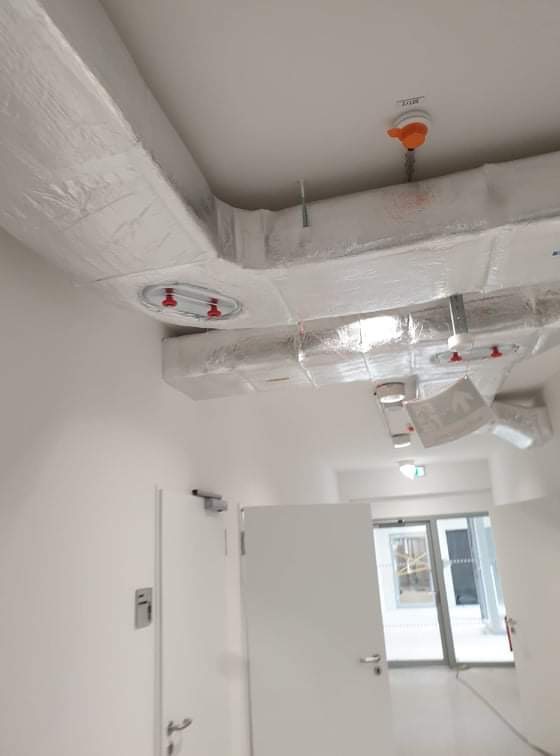 Białe kanały wentylacyjne owinięte folią ochronną, zamontowane pod sufitem w jasnym korytarzu budynku, widoczne elementy mocujące i otwory rewizyjne.