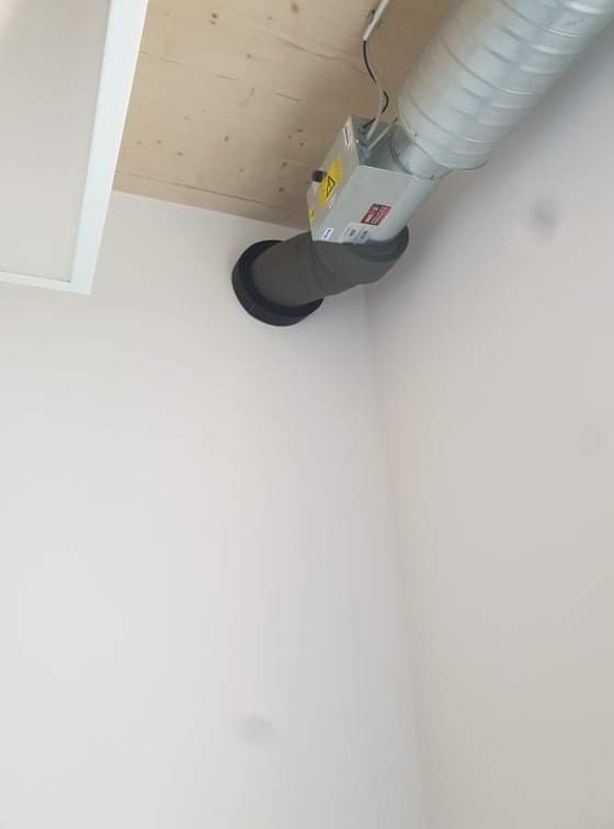 Szary przewód wentylacyjny podłączony do urządzenia z żółtą naklejką ostrzegawczą, zamocowanego do sufitu w rogu pomieszczenia z białymi ścianami.