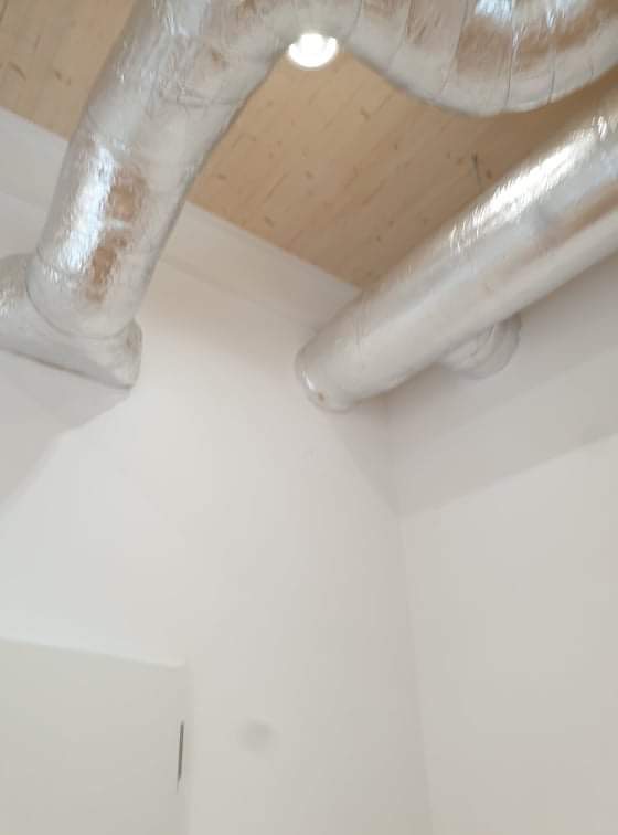 Widok od dołu na izolowane, srebrzyste rury wentylacyjne pod drewnianym sufitem w rogu białego pomieszczenia.