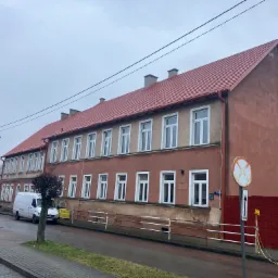 Długi budynek z nową czerwoną dachówką, widoczny z ulicy w pochmurny dzień. Na pierwszym planie znak drogowy i zaparkowany biały van.