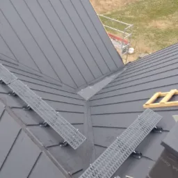 Widok z góry na fragment dachu z blachy w kolorze grafitowym, z zamontowanymi metalowymi stopniami komunikacyjnymi i elementami drewnianej konstrukcji w trakcie montażu.