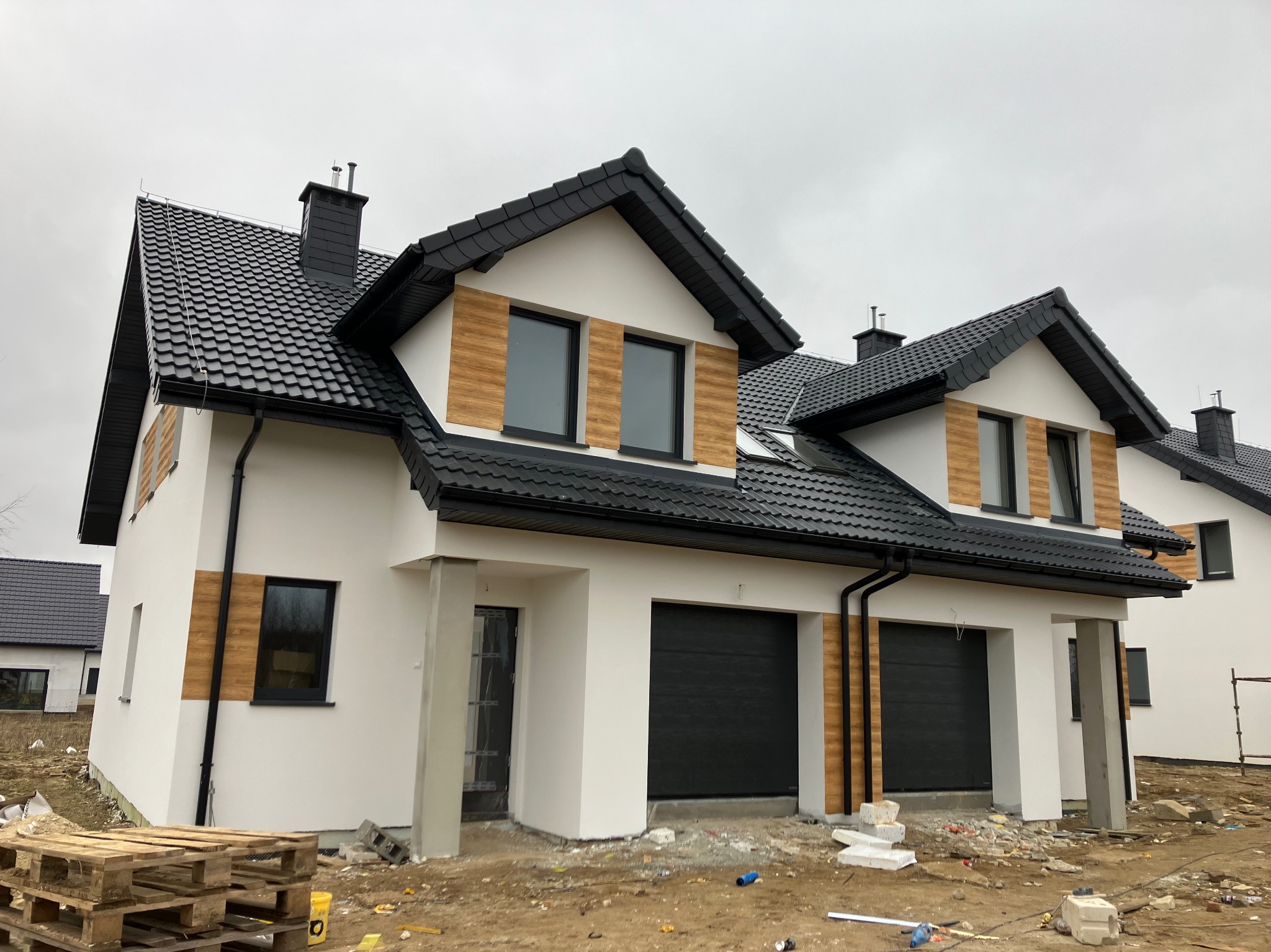 Nowoczesny, dwukondygnacyjny dom z czarną dachówką, drewnopodobną elewacją i dwoma garażami w zabudowie bliźniaczej, widok z zewnątrz na placu budowy.