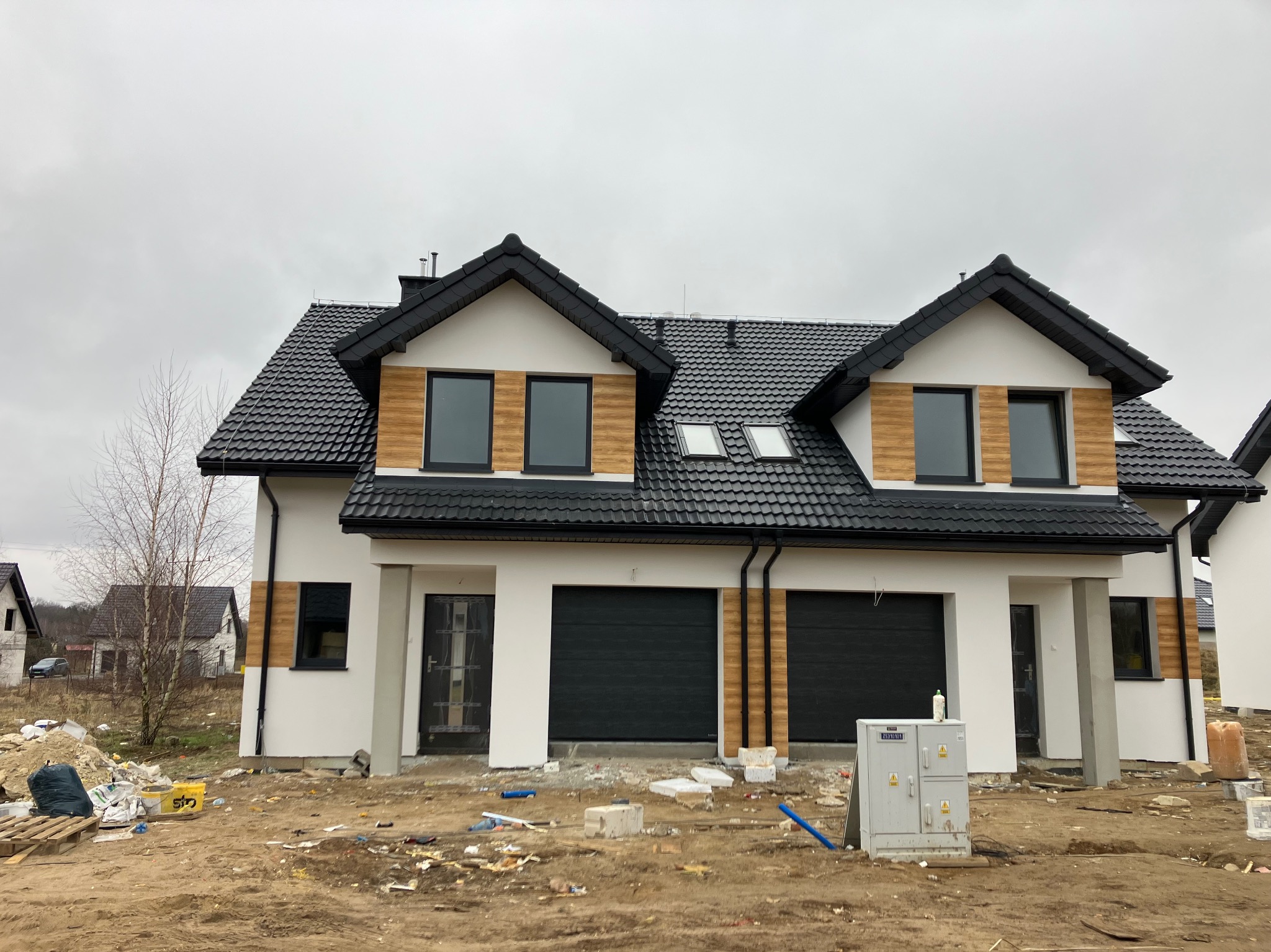 Dwukondygnacyjny bliźniak z czarną dachówką i drewnianą elewacją w trakcie wykańczania, widoczne ślady budowy i skrzynka elektryczna na pierwszym planie.