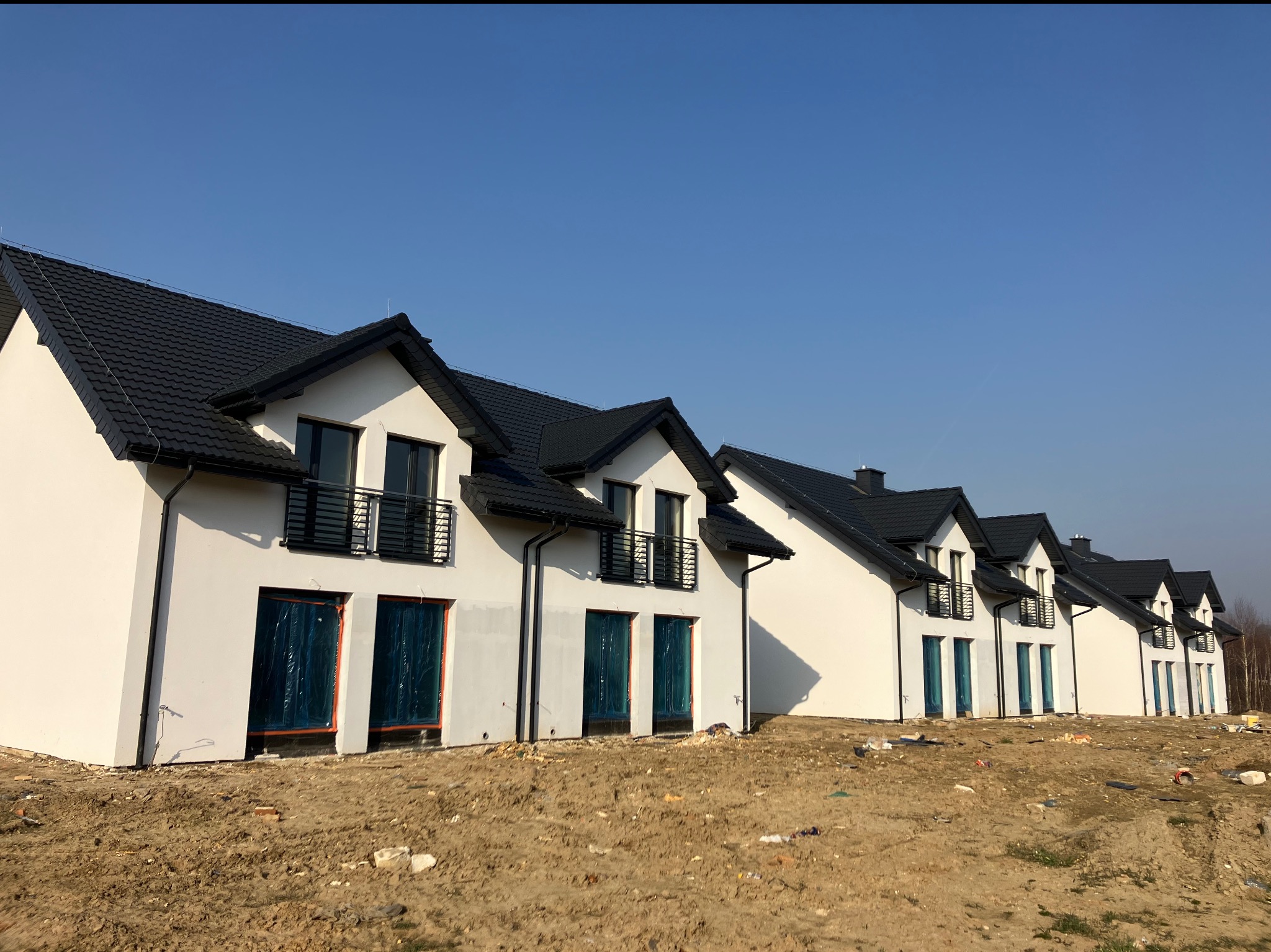 Szereg nowo wybudowanych, białych domów jednorodzinnych z czarnymi dachami i balkonami, zabezpieczone okna folią, teren wokół w stanie surowym.