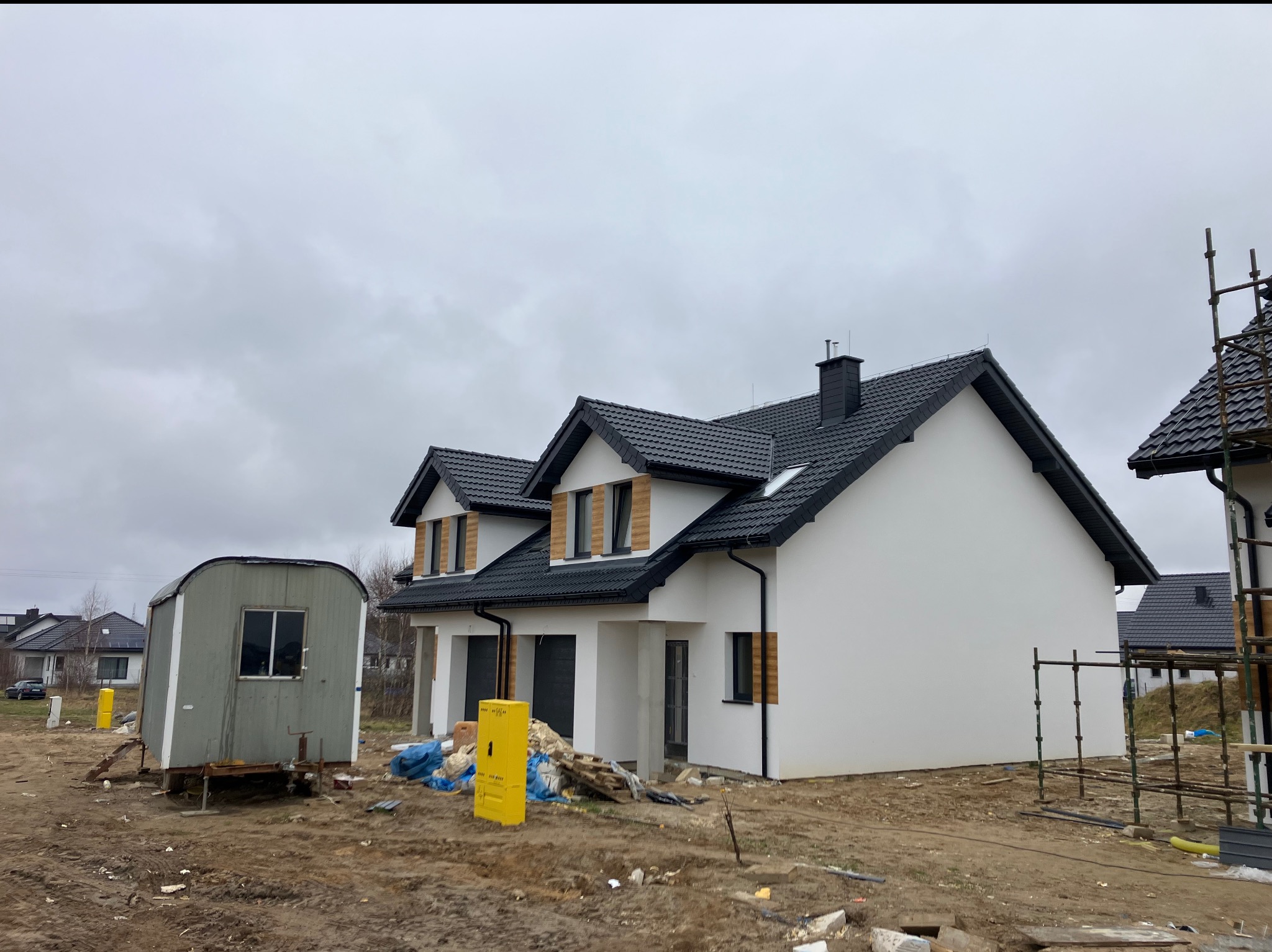 Dwa bliźniacze domy w stanie deweloperskim z czarną dachówką i białymi ścianami, widoczne rusztowania i barakowóz na działce, pochmurne niebo.