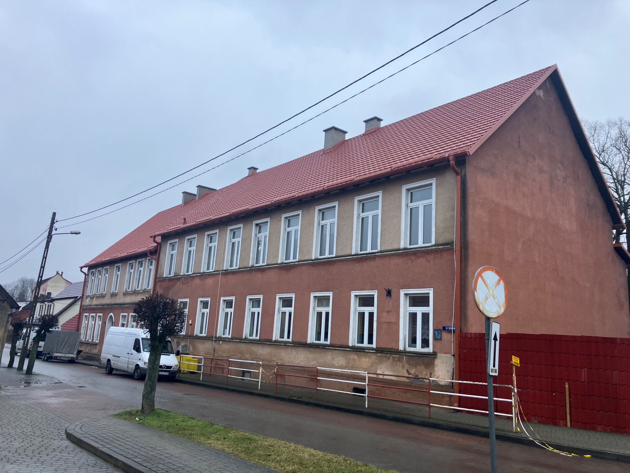 Długi budynek z nową czerwoną dachówką, widoczny z ulicy w pochmurny dzień. Na pierwszym planie znak drogowy i zaparkowany biały van.