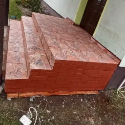 Wykończone płytkami schody wejściowe do domu, z brązowymi płytkami imitującymi cegłę na podstawie i płytkami w odcieniach brązu i beżu na stopniach, widoczne przewody elektryczne i wąż ogrodowy w tle.