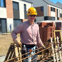 CONCRETEX GROUP SPÓŁKA Z OGRANICZONĄ ODPOWIEDZIALNOŚCIĄ - Uśmiechnięty pracownik budowlany w żółtym kasku i okularach przeciwsłonecznych, trzymający zbrojenie na tle budowanych domów i cegieł.