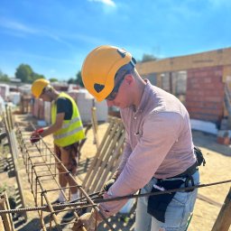 CONCRETEX GROUP SPÓŁKA Z OGRANICZONĄ ODPOWIEDZIALNOŚCIĄ - Dwóch pracowników w kaskach i roboczych ubraniach wiąże zbrojenie fundamentów na budowie domu z cegły w słoneczny dzień.
