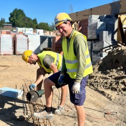 CONCRETEX GROUP SPÓŁKA Z OGRANICZONĄ ODPOWIEDZIALNOŚCIĄ - Dwóch pracowników budowlanych w żółtych kamizelkach i kaskach, jeden uśmiechnięty stoi, drugi przycina metalowe pręty zbrojeniowe szlifierką kątową na placu budowy, w tle cegły i bloczki.