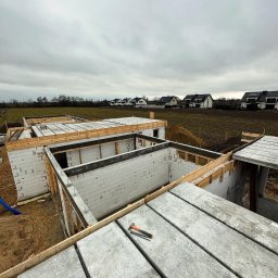 CONCRETEX GROUP SPÓŁKA Z OGRANICZONĄ ODPOWIEDZIALNOŚCIĄ - Budowa domu jednorodzinnego w trakcie wznoszenia ścian z bloczków betonowych i zalewania stropu, widoczne drewniane szalunki, zbrojenia i narzędzia na betonowych płytach stropowych, w tle zabudowa...