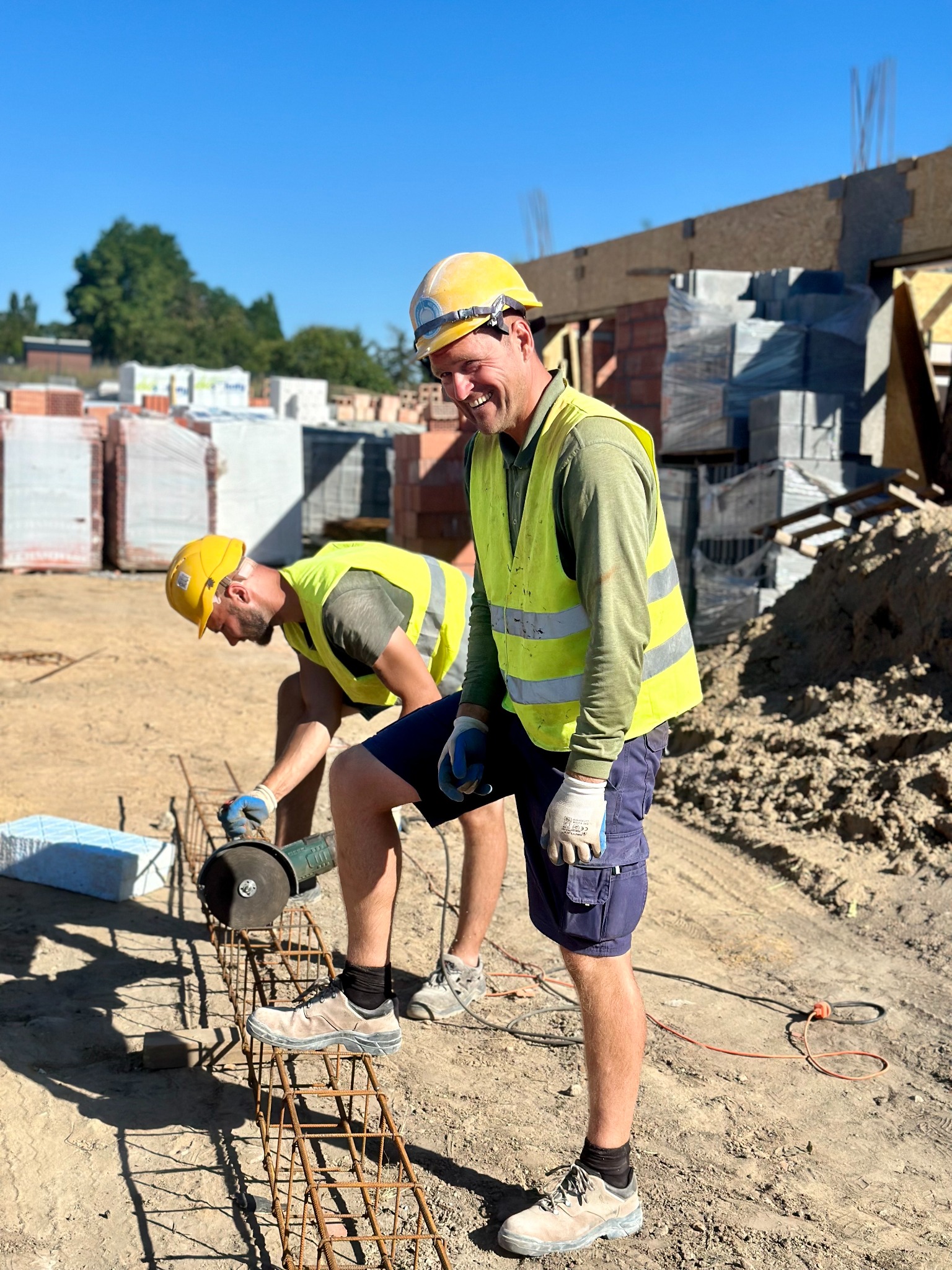 Dwóch pracowników budowlanych w żółtych kamizelkach i kaskach, jeden uśmiechnięty stoi, drugi przycina metalowe pręty zbrojeniowe szlifierką kątową na placu budowy, w tle cegły i bloczki.